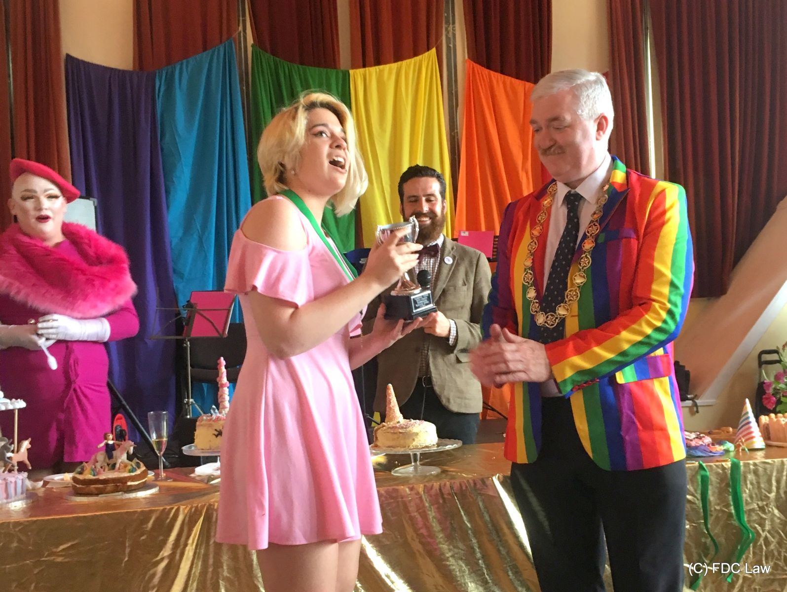 girl in pink dress accepting prize cup from a man wearing a rainbow jacket