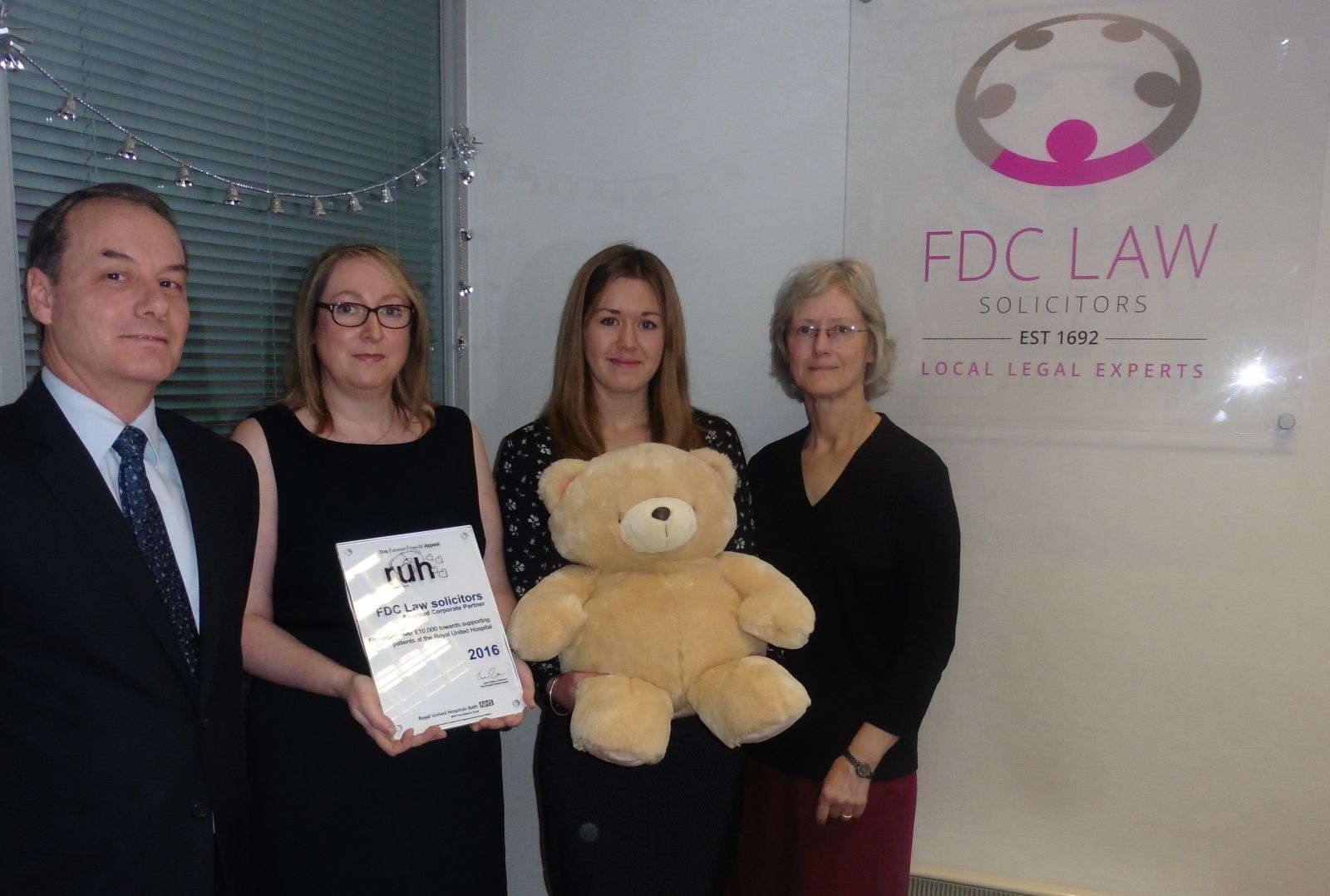Presentation of plaque to FDC Law. Group of 4 people holding palqye and teddy bear, in FDC Law office