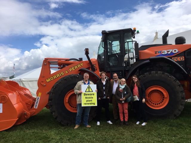 John Kilmister standing in front of a large tractor and holding a sign reading 'beware mobile plant reversing', accompanied by 4 others